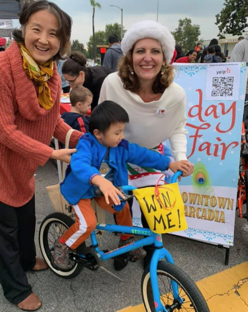 two women holding onto a young boy in a blue coat seated on a bicycle with a sign that says Win Me
