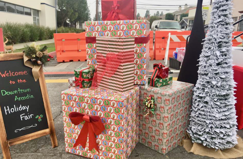 a display of wrapped packages next to a white Christmas tree and sign for Downtown Arcadia Holiday Fair