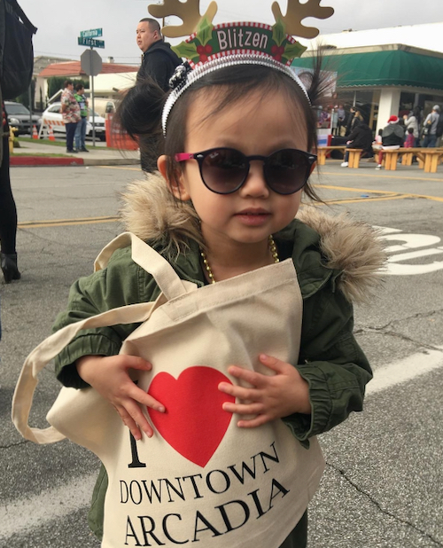 a young girl in a green jacket and reindeer antlers holding a shopping bag that says I love Downtown Arcadia