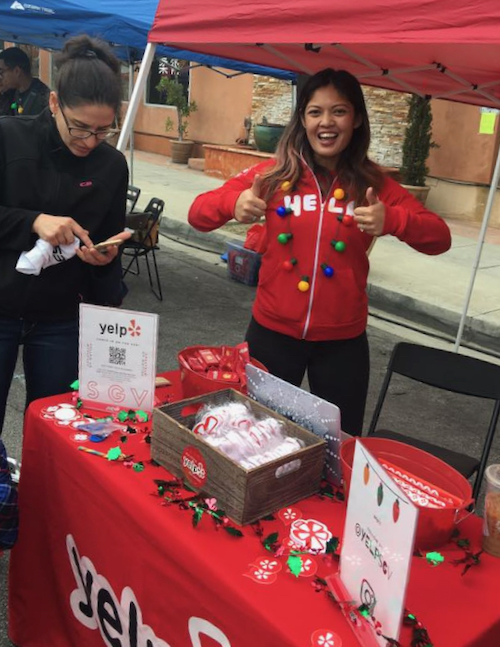 a woman in a red jacket and wearing Christmas lights as a necklace standing in a booth in front of a red table with the Yelp logo on it
